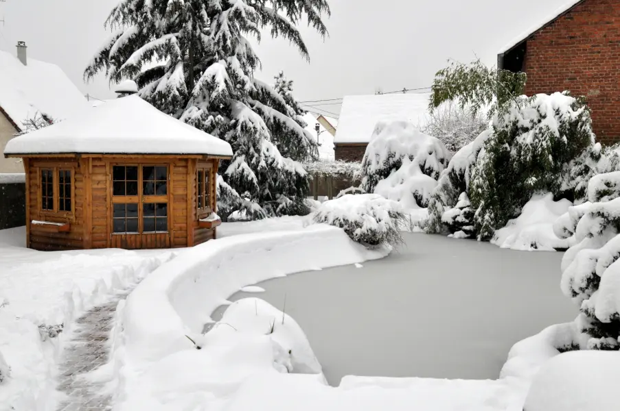 Verschneites Gartenhaus neben zugefrorenem Teich